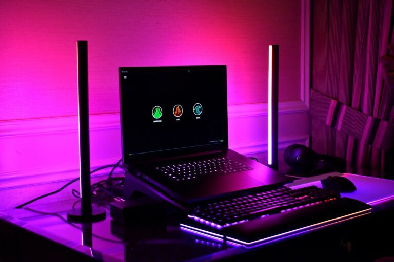 Laptop de gaming așezat pe un cooler RGB iluminat cu albastru și mov într-un setup modern